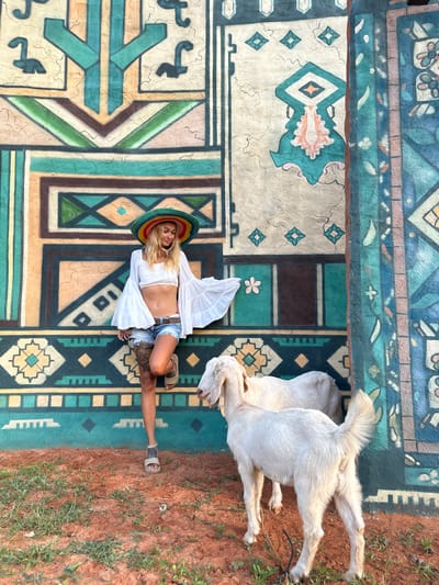 A blonde woman wearing a white crop top, denim shorts, a colorful sombrero, and sandals poses against a vibrantly painted wall with geometric patterns in Mui Ne, Vietnam. Two white goats stand in the reddish-brown dirt in front of her. The wall features a large, intricate mural in shades of teal, peach, brown, and cream, with abstract shapes and tribal-inspired motifs. The woman leans against the wall, her left arm extended, with her head tilted down. One goat is positioned in the foreground, facing away from the camera, while the other is partially visible behind it. The ground is covered with sparse green vegetation.