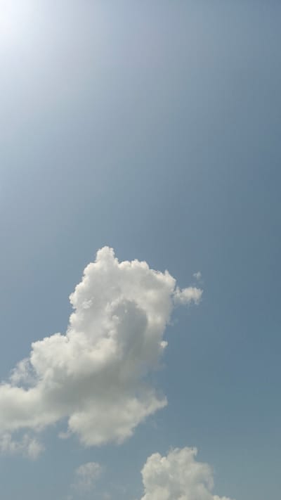 The image depicts a clear blue sky with a large, fluffy white cumulus cloud dominating the lower center. A faint bright light, possibly the sun, is visible in the upper left corner, casting a soft glow on the sky. The lower portion of the image shows hints of more scattered clouds. The scene is a natural outdoor environment, with no signs of human presence or urban elements, including the city of Serti, Nigeria.