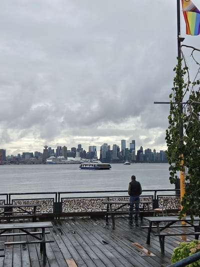 The image shows a view of the Vancouver skyline from North Vancouver, Canada, on a cloudy day. In the foreground, a wooden pier with several picnic tables is wet, indicating recent rain. A man stands on the pier, facing away from the camera, looking out at the water. A fence behind the man is covered in numerous colorful padlocks. In the middle ground, the Burrard Inlet separates North Vancouver from the city. Several ferries and a large cruise ship are visible on the water. The Vancouver skyline dominates the background, with numerous high-rise buildings and the Vancouver Lookout tower visible. A rainbow pride flag is flying on a pole to the right of the image. A tree with green leaves hangs into the frame from the right side.