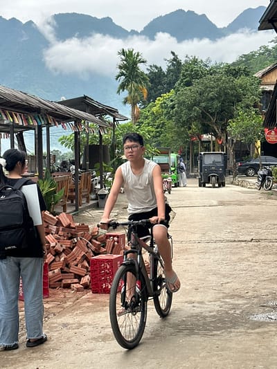 In Mai Chau, Vietnam, a young man wearing a white tank top and black shorts cycles down a dusty, rural road, his right foot in a flip-flop pedaling. Behind him, a young woman with a black backpack on her shoulders, wearing a white and black t-shirt and light blue jeans, stands facing away from the camera. To their left, a large pile of red bricks and red crates are stacked on the side of the road. In the background, a traditional wooden building with a thatched roof, adorned with colorful flags, stands near lush green trees and the misty peaks of mountains characteristic of the region. Several vehicles, including a black tuk-tuk and a green passenger van, are visible further down the road, suggesting a bustling but unpaved commercial or residential area. The sky is overcast, with clouds partially obscuring the mountain range.