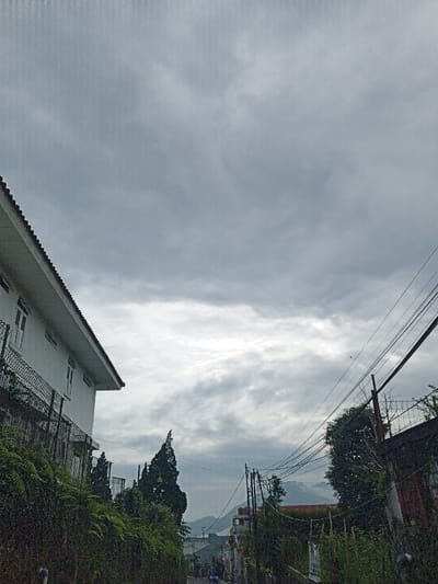 A cloudy sky fills the frame, looming over a downward sloping street. On the left, a white building with a red-tiled roof is partially visible, separated from the road by a chain-link fence and lush vegetation. Power lines crisscross the sky on the right, running alongside greenery and a building with a textured wall. In the distance, hills can be seen through a break in the clouds, hinting at a village or town. The scene evokes a quiet, overcast day in a hillside community.