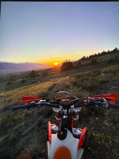 This media file captures a sunset over a mountainous landscape, with a dirt bike prominently featured in the foreground. The image is taken from a slightly elevated perspective, looking down and across the scene.
The main subject is an orange and white dirt bike, positioned on the right side of the frame, angled towards the left. The handlebars, front forks, parts of the engine, the white seat, and the orange rear fender are clearly visible. The handguards are orange, and the brand "KTM" is discernible on one of them. The "WP" logo, indicating WP Suspension components, can be seen on parts of the front fork assembly. The bike is parked on uneven, grassy terrain dotted with small yellow flowers.
In the background, a vast panorama unfolds, dominated by a setting sun on the horizon. The sun's intense orange glow illuminates the sky and casts a warm hue over the distant mountains and valleys. The sky transitions from a bright orange near the sun to softer yellows and blues higher up. A faint white contrail from an aircraft is visible in the upper left portion of the sky. The landscape consists of rolling hills and mountains, covered in dark green vegetation, likely forests or fields. The scene suggests a rural or wilderness setting, fitting the provided location context of Razlog, Bulgaria, which is surrounded by mountain ranges like Pirin and Rhodope.
The time of day is clearly sunset, indicated by the sun's low position and the long shadows, creating a serene and picturesque atmosphere. The weather appears clear and calm. The dirt bike is stationary, suggesting a pause during an off-road ride to enjoy the scenic view, or the end of a day's adventure. The image exhibits visible horizontal scanning lines, indicating it was likely a photograph taken of a digital screen or monitor.
