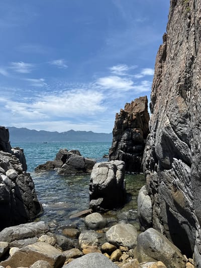 The image depicts a rocky coastline in Vĩnh Lương, Vietnam, under a clear blue sky with scattered white clouds. Large, rugged grey and brown rocks dominate the foreground and midground, some partially submerged in the turquoise ocean water. The water is clear, revealing smaller rocks beneath the surface. In the distance, a range of hazy blue mountains forms the horizon. A small boat can be seen on the water. The scene is devoid of any people.