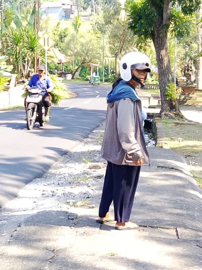 In Tabanan, Indonesia, a woman stands on a paved sidewalk next to a road. She wears a white helmet, a grey hooded jacket over a light blue shirt, and dark blue pants. In the background, a man rides a motorcycle carrying bundles of green foliage. The scene is illuminated by sunlight, casting shadows on the road and sidewalk. Lush green vegetation and trees are visible, along with buildings and a small green cart in the distance.