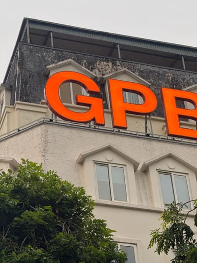 The image shows a close-up, low-angle view of the top of a building in Hanoi, Vietnam. Large, bright orange neon letters spelling "GPB" are prominently displayed across the facade. The letters are mounted on a dark, textured wall that appears aged or weathered. Below the letters, there are several dormer windows with white frames and light-colored roofs. Lush green foliage from trees obscures some of the lower parts of the building and the foreground. The sky above is overcast.
