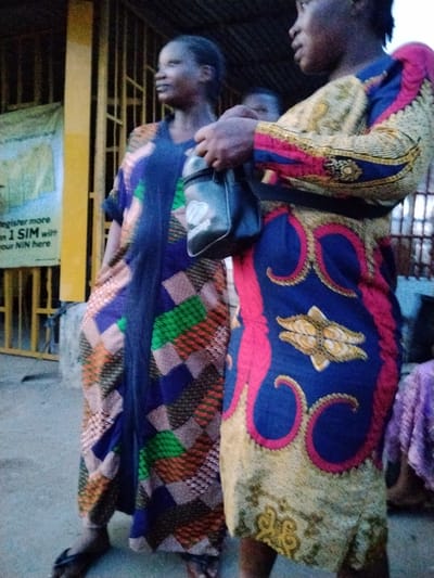 In Ura Nta Umuarandu, Nigeria, two women stand close to each other on what appears to be a street. The woman on the left is wearing a floor-length gown patterned with blue, green, and orange geometric shapes. She looks off to the left. The woman on the right wears a patterned dress of yellow, blue, and pink, and a black bag is slung over her shoulder. The background includes a building with metal bars and a sign, and there are other people further back in the scene.
