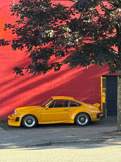 A yellow Porsche 911 Carrera is parked on the side of a street, parallel to a red brick building. The car is facing towards the right of the frame. A dark-colored tree with sparse, dark leaves casts shadows across the building and the car. The building has a dark door with a yellow metal gate to its right. The street is paved, and there is a sidewalk in front of the building. The scene appears to be a daytime shot, with sunlight creating distinct shadows. The location is not specified in the image or crops.