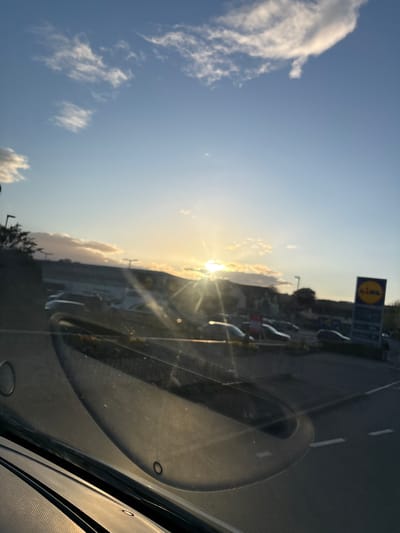 The image, taken from inside a vehicle, shows the sun shining over what appears to be a parking lot and street in Oswestry, United Kingdom. A Lidl sign is visible amidst parked vehicles. The sky is blue with scattered clouds. The interior of the car is visible in the foreground.
