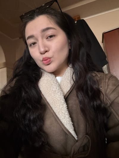 A woman with dark hair is wearing a brown jacket with a white, sherpa-lined collar. She has brown eyes and is pouting her lips. She is also wearing glasses. A black bag is behind her. The location is unknown and cannot be discerned from the image.