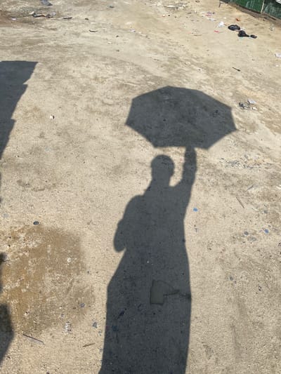 In Iranla, Nigeria, a shadow of a person holding an umbrella is cast on a dirty, paved area. The ground is covered in debris, including plastic bags, wrappers, and other litter. The shadow of the person is elongated and distorted by the low angle of the sun, obscuring details of their features. The umbrella's shadow is clearly visible above the person's head. The environment appears to be an outdoor space, possibly a street or a vacant lot, that is not well-maintained.