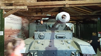The image shows a Panzerkampfwagen IV tank on display at the Yambol Military Museum in Yambol, Bulgaria. The tank is positioned indoors under a wooden roof structure. Two mannequins in German military uniforms are seated in the tank. A sign next to the tank has text in Bulgarian and English explaining that the tank was designed to break enemy defenses and destroy infantry and weapons. People can be seen to the side of the tank and partially blurred in the foreground. A fire extinguisher is visible on the wall.