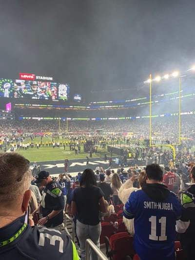 A packed stadium at night with Levi's Stadium visible. Spectators watch the event, some wearing Seattle Seahawks jerseys. One jersey displays "SMITH-NJIGBA" and the number "11." Another jersey shows the number "31." LED displays line the stadium, adding to the bright atmosphere. People are sitting in the stands.