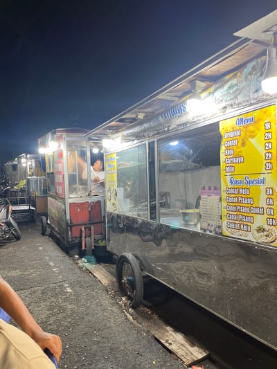 This media file shows a street food vendor stall at night in Banda Aceh, Indonesia. The stall is illuminated by bright lights, highlighting a menu board advertising various food items like "Original," "Coklat" (chocolate), "Keju" (cheese), "Sarikaya," and "Milo" under the "Menu" section. A "Rasa Spesial" (Special Flavor) section lists "Coklat Keju" (chocolate cheese), "Canai Pisang" (banana flatbread), and "Canai Pisang Coklat" (chocolate banana flatbread) with prices. The vendor, a person wearing a white shirt, is visible working inside the stall behind a glass partition. In the background, other food stalls and a motorcycle are present, indicating a busy street food scene. The ground appears to be asphalt, and there are some discarded items on the side of the road. The overall impression is of a lively night market or street food area.