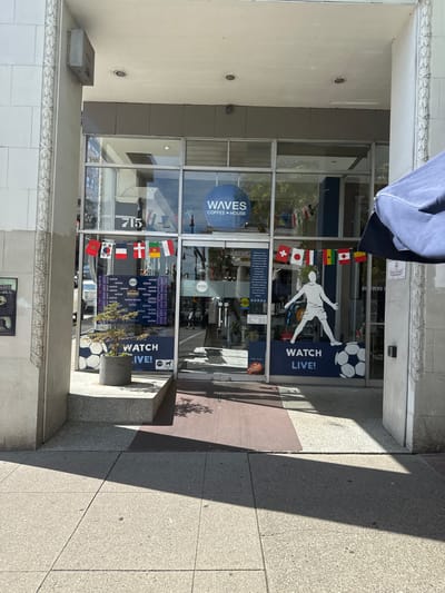 The image shows the exterior of a coffee shop named "WAVES COFFEE HOUSE" located at 715 Carnarvon Street in New Westminster, Canada. The shop has large glass windows displaying flags of various countries, a large screen showing a grid, a silhouette of a soccer player next to a soccer ball, and the text "WATCH LIVE!". A small potted tree is placed in front of the window. The entrance to the shop is visible, with a ramp and a mat leading up to it. The building's facade is made of concrete with decorative elements. The sidewalk in front of the shop is made of concrete tiles. Shadows from the building and overhead structures are cast on the sidewalk and the entrance. The lighting suggests it is daytime with bright sunlight. No people are clearly visible within the shop, but a few figures are partially seen through the glass.