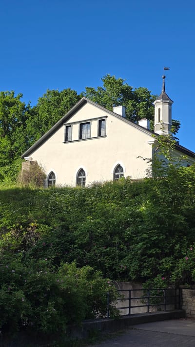 This is a vertical, outdoor, daytime photograph of a light-colored building with a dark roof, possibly a church or historical structure, in Keila-Joa alevik, Estonia. The building features a tower with a weather vane on top. The structure has several windows, some rectangular and others arched. Abundant green trees and foliage surround the building, with some smaller pink flowers visible among the greenery. To the right of the building, a dark metal fence and a paved path are visible. The sky is clear and bright blue, suggesting fair weather. The overall impression is one of serene natural beauty and historical architecture. No people are visible in the image, and no text is present.