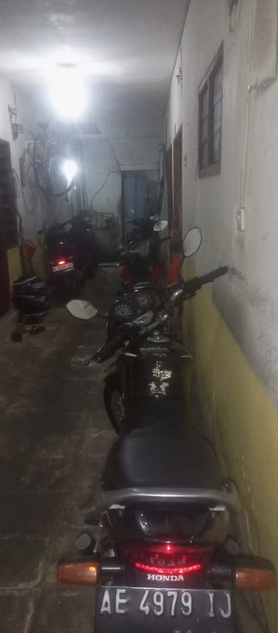 The image depicts a narrow, dimly lit passageway in a residential building in Kecamatan Waru, Indonesia, at coordinates -7.35 latitude and 112.764 longitude. The passageway appears to be a garage or carport. It is lined with whitewashed walls, with a yellow patch near the lower right corner. A black motorcycle is parked in the foreground, facing away from the camera. The motorcycle has a black seat, a black rear fender, a red taillight, a license plate reading "AE 4979 IJ", and a "Honda" emblem on the rear. The motorcycle is partially obscured by the wall on the right side of the image. A black car with a red taillight and a license plate reading "W 5066 VJ" is parked behind the motorcycle. The car is partially obscured by the wall on the left side of the image. In the upper right corner of the image, a window can be seen, with a white frame and a brown windowpane. In the upper left corner, the outline of a bicycle can be seen, but the details are obscured. A small light fixture hangs from the ceiling in the center of the image, illuminating the scene with a soft, white glow.