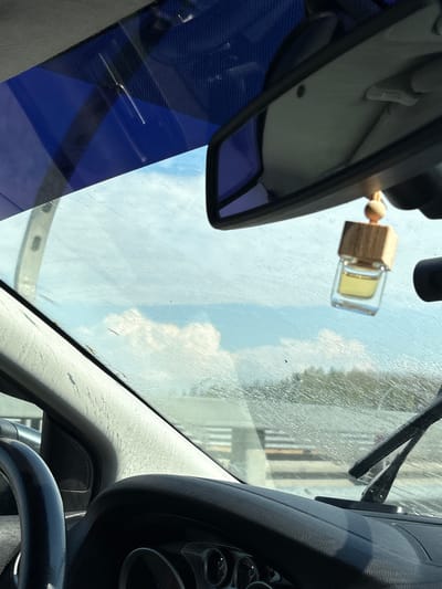 The image shows a view from inside a car, likely stationary, through the windshield. The sky with white clouds is visible through the windshield. A rearview mirror and a small, wooden-topped air freshener are hanging from the top of the windshield. A blue-tinted section of the windshield is visible on the lower left. A windshield wiper is present on the right side. Part of the car's dashboard, including the steering wheel and instrument panel, is in the lower right. A building is vaguely visible through the dirty windshield. The location is indicated as Beloostrov, Russia.