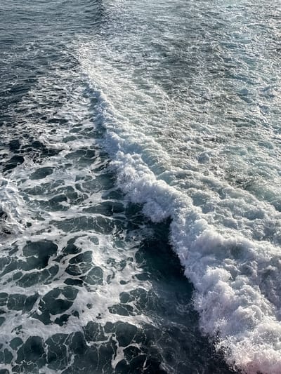 The image displays a close-up, high-angle view of agitated ocean water, likely a vessel's wake, in the marine environment near Kamariotissa, Greece. The dominant feature is a diagonal band of churning, white foam and disturbed water extending from the lower right to the upper left. This foamy trail is approximately one-third the width of the frame at its widest point, with a defined leading edge forming a small wave crest. To the left of this primary disturbance, the water appears darker, a deep teal-blue, interspersed with irregular patches of white foam. To the right, the water is also turbulent but appears less defined, with widespread, finer white foam and ripples covering a grey-blue surface. No specific objects or individuals are visible beyond the water itself and its surface patterns. The scene suggests movement through the water, generating the depicted wake.