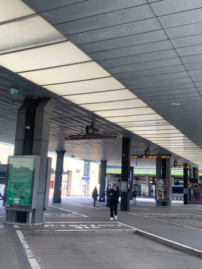 The image shows the interior of a bus station in Kraków, Poland, located at latitude 50.068 and longitude 19.949. It's a covered waiting area with a grey concrete ceiling and several pillars supporting the roof. The floor is made of grey concrete, with white painted markings. There is a sign on a pole that reads "G4 G1n," and another sign reads "G4." There are a few people waiting in the area, including one woman who is wearing a black jacket and black pants and is holding a phone in her left hand. There are several large posters on the walls, and a few bins with garbage bags in the background. Several bus bays are visible, and the bus station is lined with trees on the outside. The sun is shining brightly.