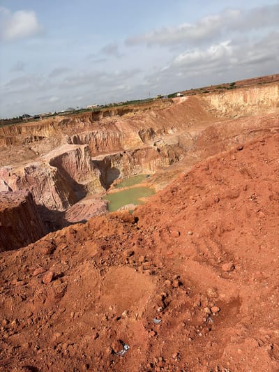 The image shows a quarry located in Kwang, Nigeria. The quarry is an open pit with steep, terraced walls of reddish-brown soil and rock. A small pool of murky green water is visible at the bottom of the pit. In the distance, there are a few buildings and vehicles, suggesting ongoing construction or mining operations. The sky is cloudy, with patches of blue visible. The foreground of the image is dominated by a pile of reddish-brown soil.