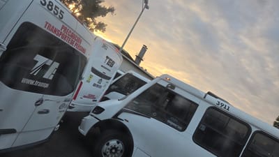 The image shows three white vehicles parked outside on a street with a utility pole and a tree in the background, silhouetted against the sky at either dusk or dawn. The vehicle in the foreground is a van with the number "3855" and the words "TRANSPORTE DE PERSONAL" printed on its side. The logo "TEB" and the website "www.transp" are visible. The second vehicle appears to be a van as well, also with "TEB," a phone number, and the number "3912". The largest vehicle is a bus with the number "3911" on its side. The location is suggestive of La Tijera, Mexico, based on the Spanish text on the vans and buses.