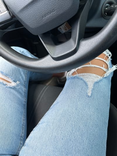 This media file is a close-up shot taken inside a vehicle, focusing on the steering wheel and the driver's legs. The driver is wearing light blue denim jeans with ripped detailing on the knees, revealing skin underneath. The steering wheel is black and textured, with "AIRBAG" clearly visible on the driver's side. The lighting suggests it is daytime, possibly sunny, given the bright exposure of the jeans. The overall impression is casual and relaxed, perhaps indicating a journey or a moment of rest during a drive. The setting appears to be a modern car interior. There are no other people or significant background details visible. The visible text is limited to the word "AIRBAG" on the steering wheel. The location context provided is Kableshkovo, Bulgaria, though no specific visual cues within the image directly confirm this location.