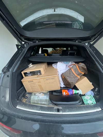 The open trunk of a dark-colored vehicle is filled with various items. There are cardboard boxes, one labeled "ILVA," a Louis Vuitton bag, a bottle of cleaning solution, and a glass jar. A green package of tissues is also visible, along with some white fabric or clothing. The car's taillight is partially visible on the right side of the frame. The contents are tightly packed within the trunk space.