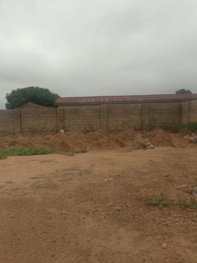 The image shows a construction site in Minna, Nigeria, under a cloudy sky. A brick wall runs horizontally across the mid-ground, with piles of excavated dirt in front of it. A building with a red roof is partially visible behind the wall. The foreground is a dirt and gravel area.