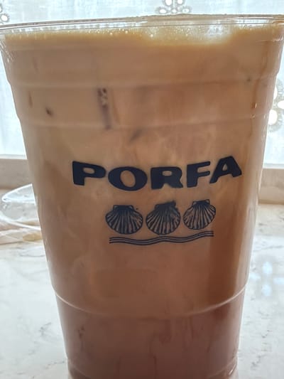 The image shows a close-up of a clear plastic cup filled with a light brown, milky beverage with ice cubes. The cup has a dark blue logo printed on it. The logo consists of the word "PORFA" in a stylized, bold font, with three scallop shells and wavy lines placed to the right of the text. The drink appears to be an iced coffee or latte. The background is out of focus but suggests an indoor setting with natural light coming from a window, possibly a cafe or home. The overall lighting is soft and diffused, indicating it could be daytime. There are no people visible in the image. The texture of the plastic cup and the condensation on the outside are visible details. The location context is Fort Lauderdale, United States of America.