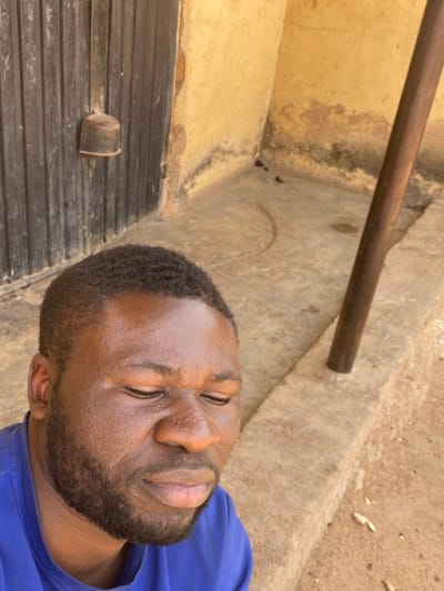 A dark-skinned male with a short beard and short, wavy dark hair, wearing a blue t-shirt, is depicted in a medium close-up, outdoors in Udu, Nigeria. His eyes are closed, and his face shows a slight grimace, suggesting discomfort or exposure to bright light. He is positioned in the lower-left foreground, looking downwards and to his right. The background features a weathered, light yellow-brown wall with dark stains and discoloration along its base. To the left, a dark, vertically paneled wooden door with a cylindrical, possibly metallic, fixture is visible. To the right, a dark brown vertical pole extends from the mid-ground upwards. The ground is a light-colored concrete or cement surface, showing a distinct semicircular groove and some small debris. The scene is illuminated by strong, direct sunlight.