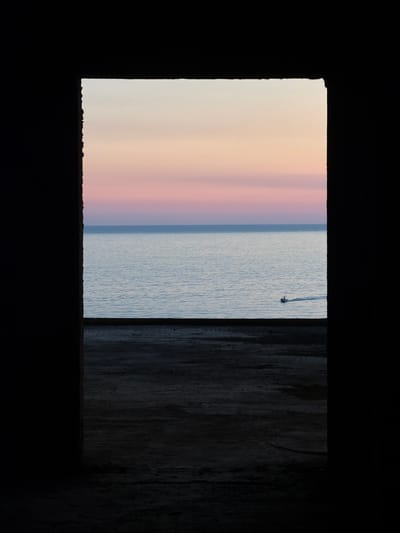 The image, taken in Perazića Do, Montenegro, presents a view of the sea and sky framed by a dark, rough opening. The opening appears to be a window or doorway of an unfinished or abandoned structure, with its edges showing exposed concrete or stone.
The scene beyond the opening is a serene seascape at sunset. The water is a calm, pale blue, reflecting the soft colors of the sky. The sky itself transitions from a light yellow at the horizon to delicate shades of pink and purple higher up. A small motorboat with two visible occupants is moving across the water, leaving a white wake behind it. In the foreground, within the dark frame, is a rough, dark ground surface, suggesting a pathway or floor made of earth and debris. The overall impression is one of quiet contemplation, with the natural beauty of the sunset contrasted against the stark, man-made opening.