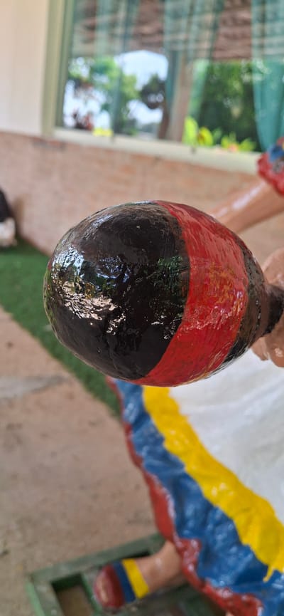 This image focuses on a close-up view of a decorative object, possibly a statue or a cultural artifact, with a glossy, textured surface. The object is predominantly black, with a prominent band of red encircling it. The colors appear vibrant and somewhat reflective, suggesting a wet or varnished finish. In the background, hints of other colors and shapes suggest the object is part of a larger display or scene. The background also shows glimpses of what could be greenery and outdoor elements, possibly a garden or a patio area, illuminated by natural light. The overall impression is that of a brightly colored, possibly festive, decorative item. The provided location context, Vallecito, Venezuela, suggests a tropical or sub-tropical environment. There is no visible text in the image.