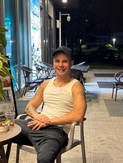 A man wearing a white tank top and a black baseball cap is sitting outdoors at night, with his legs crossed. He has tattoos on his arm and is smiling at the camera. A table with a drink and some colorful objects is in the foreground, and chairs are visible in the background. Behind the man, there is a large illuminated sign with what appears to be Chinese text and images of cars. The setting suggests an urban environment in Huangpu District, China, possibly a cafe or restaurant patio. The lighting indicates it is nighttime.