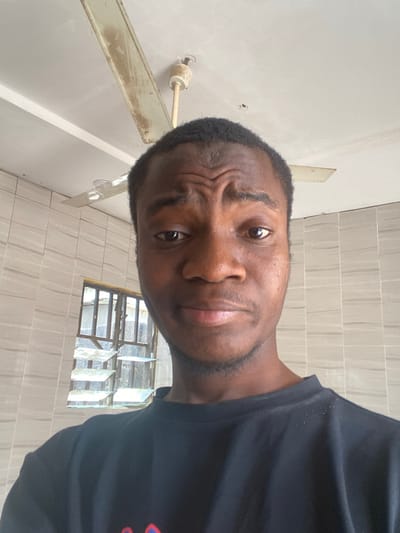A close-up, head-and-shoulders indoor photograph depicts a dark-skinned male individual with short, dark hair, looking directly at the camera. His eyebrows are slightly furrowed, and his mouth is in a neutral or subtly smirking expression. He wears a black short-sleeved t-shirt. The background features a light-colored ceiling with a visible, discolored ceiling fan possessing at least two blades. The walls are covered with light-colored, horizontally arranged rectangular tiles. To the viewer's left, a window with multiple louvered glass panes and a metal frame offers a view of an indistinct exterior. This scene is situated within a room in Minna, Nigeria.