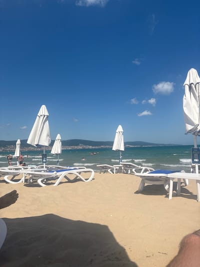 The media file captures a sunny day at a beach in Nesebar, Bulgaria. The scene is dominated by a bright blue sky with a few scattered white clouds. In the foreground, there is a sandy beach with several white lounge chairs and closed white umbrellas lined up. The chairs cast long shadows on the sand, indicating it is likely midday. The sea in the background is a vibrant green, with moderate waves breaking on the shore. A distant coastline with buildings and hills is visible across the water, suggesting a populated area. Some people can be seen in the water and on the beach, enjoying the sunny weather. One of the umbrellas has a small sign with text, including a QR code and numbers "4 3". The overall impression is one of a relaxed summer day at a popular beach resort.