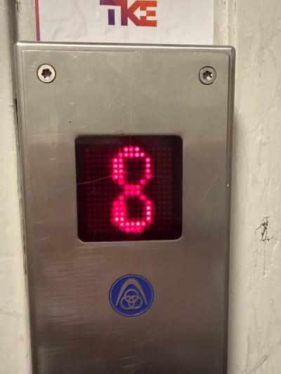 An elevator control panel displays the number "8" in bright pink LED lights. The panel is made of brushed metal and has two star-shaped screw heads visible at the top. Below the number display, a circular blue emblem with a stylized design is present. The panel is mounted on a textured, off-white wall. A partial view of a sign with the letters "TKE" in purple and orange is visible at the top of the frame. The scene depicts a functional elevator indicator, possibly located in a public building or transportation hub in Oslo, Norway.