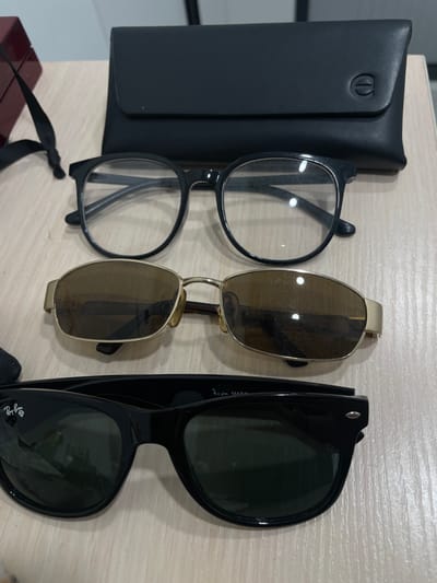 The image shows a close-up, overhead view of three pairs of glasses and a black case on a wooden surface. The glasses are stacked vertically. The top pair is black-rimmed prescription glasses. The middle pair has gold-toned rims and brown lenses. The bottom pair is black Wayfarer-style sunglasses with a Ray-Ban logo on the left lens. The black case is behind the glasses. The scene is indoors, likely on a table or desk. The specific location of Kommunarka, Russia, is not visually discernible from the image.