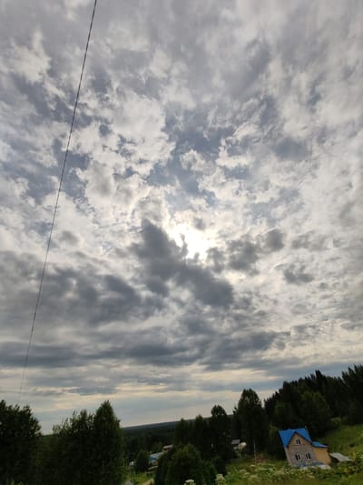 A wide-angle, high-angle shot captures a cloudy sky with the sun peeking through the clouds on the right side of the frame. A thin black wire extends diagonally from the top left to the bottom left of the image. Below the sky, a dense line of green trees stretches across the middle ground. On the right side of the frame, a small house with a bright blue roof and a beige exterior is visible nestled among the trees. The house has several windows. In the foreground, there are green trees and some white wildflowers. The landscape appears to be rural. This image was taken in Chukhlem, Russia.