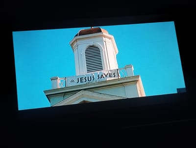 The image depicts a church steeple against a clear blue sky. A banner reading "JESUS SAVES ROMANS 10:13" is draped across the steeple's railing. The steeple is white with a brown roof. The image appears to be taken from a low angle. There is no evidence in the image to suggest this scene is located in Punta de Piedras, Venezuela. Therefore, I cannot comment on the location.
