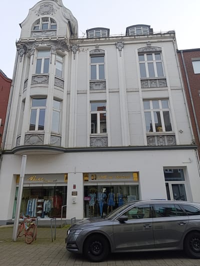 The image presents a street scene in Emmerich on the Rhine, Germany. The focus is on a multi-story building with architectural details and a storefront on the ground floor. The building's facade is predominantly light-colored, with ornate window designs. On the ground floor, there are two storefronts: "Butik Emmerich" and "LIVE on Facebook" signage. A gray car is parked in front of the building, partially obscuring the storefronts. A red bicycle is parked near the entrance of "Butik Emmerich." The sidewalk is paved with interlocking blocks. The sky is overcast.