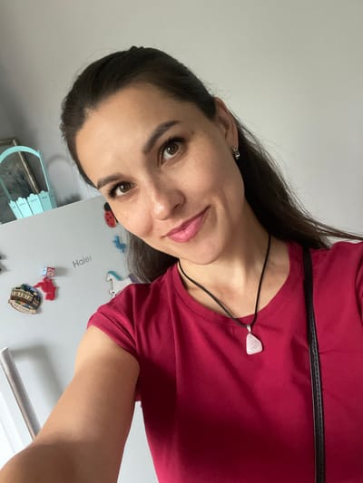 A medium shot shows a woman with dark hair and brown eyes taking a selfie in front of a refrigerator. She is wearing a red t-shirt and a necklace with a light pink pendant. The refrigerator has several magnets on it, including a ladybug, a blue bird, a unicorn, and a magnet with text that appears to be in Russian. A light blue basket with a decorative pattern is visible on top of the refrigerator. The background is a plain grey wall. The woman's right arm is extended, holding the phone. The overall lighting is bright and natural. There are no clear indicators of the specific location within Donskoye, Russia, as the image is taken indoors with a plain background.
