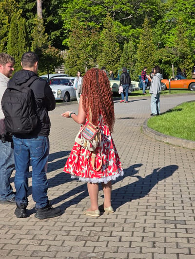 The image shows a sunny outdoor scene in Repino, Russia, where a group of people are viewing classic cars. In the foreground, a woman with long braided red hair, wearing a red and white patterned dress with white lace trim, tan stockings, and gold shoes, stands on a paved walkway. She has a small, colorful bag on her shoulder. A man wearing jeans, a black jacket, and a backpack stands next to her. The paved walkway is bordered by a grassy area. Further in the background, various classic cars of different colors are visible, along with more people standing and looking at the cars. The presence of trees suggests the area might be near a park or a wooded area. The shadows suggest a sunny day.