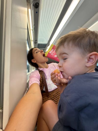 In an interior shot of a train carriage, a woman in a pink long-sleeved shirt with a leopard print strap across her chest is captured mid-action as she takes a bite from a bag of chips. To her right, a young child in a blue shirt is being offered a snack by an unseen person holding out a piece of food. The child is looking at the snack intently. The train carriage interior features overhead lighting and ribbed walls. The overall lighting suggests daytime. There is no visible indication that this scene is in Tarragona, Spain.