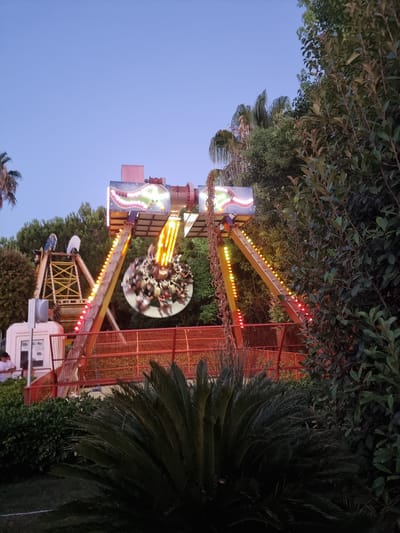 This is a medium-shot, eye-level, vertical photograph of an amusement park ride in Manavgat, Turkey. The ride, a swinging pendulum attraction, is centrally located and in motion, with a circular gondola filled with riders spinning. The ride is brightly lit with yellow and red lights along its support beams, and the top of the structure has a colorful, illuminated sign. To the left of the main ride, a yellow metal structure with a crane-like element is visible, and a white booth with a person standing in front of it is partially obscured by the structure. The background consists of lush green trees, including palm trees, and a clear, light blue sky. In the foreground, a large, dark green palm-like plant with spiky fronds is prominent. The overall impression is of a lively, evening scene at an amusement park.