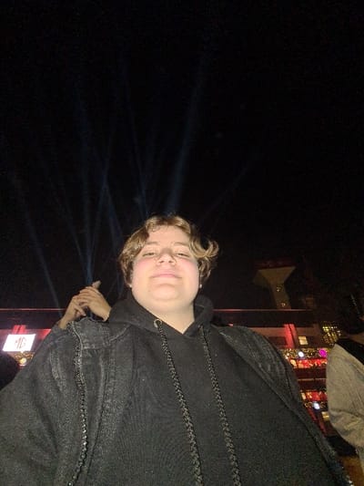A person with blondish hair and a fair complexion is wearing a dark hoodie and denim jacket. The background is dark with faint laser beams extending into the darkness. A building with brightly lit windows and a white spotlight are visible in the upper right corner. The subject appears to be outside at night, possibly at an event.