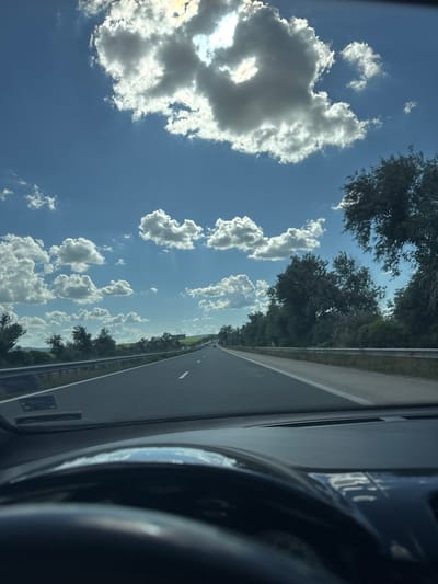 This media file captures a view from inside a vehicle traveling along a highway in Gabarnitsa, Bulgaria. The scene is dominated by a bright blue sky filled with fluffy white clouds, suggesting a clear and sunny day, likely midday due to the strong sunlight. The road stretches ahead, bordered by a line of trees on the right side, with more trees visible in the distance. The perspective from inside the car includes a portion of the steering wheel and dashboard on the right, indicating the forward direction of travel. The overall impression is one of a pleasant journey through a rural or semi-rural landscape. There is no visible text in the image.