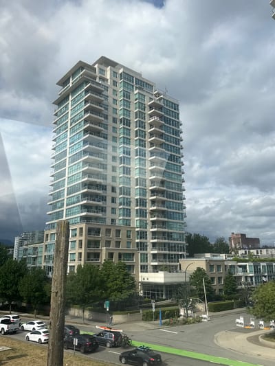 The image captures a view of the residential buildings and streets in Vancouver, Canada. A tall, multi-story condominium building with balconies and numerous windows dominates the central part of the frame. The building has a modern architectural style, with light-colored facade and blue-tinted windows. In front of the condominium, there are several other residential buildings and lush green trees. The street below is populated with various vehicles, including cars, a truck, and a SUV carrying bicycles and a kayak on its roof. A pedestrian can be seen walking on the sidewalk. The sky is filled with dense, gray clouds, suggesting an overcast day. A wooden utility pole stands in the foreground, partially obscuring the view.