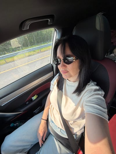 In the city of Fyodorovo, Russia, a woman with dark hair and sunglasses is in the passenger seat of a car, taking a selfie. She is wearing a light-colored t-shirt and light-wash jeans. A seatbelt is visible across her chest. Through the car window, a road with a yellow line and guardrail is visible, bordered by trees. The car's interior, including a door handle and a small green toy on the door panel, is also visible.