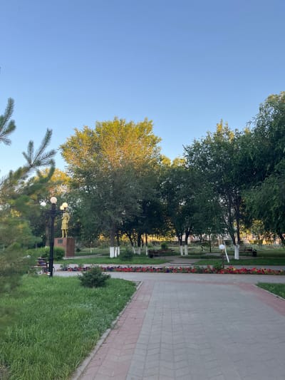 The image depicts a park scene in Qobda, Kazakhstan. The setting is an outdoor park with a paved walkway lined with grass and flowerbeds. Tall trees with lush green foliage dominate the background, with some of their leaves catching the sunlight, suggesting it is either early morning or late afternoon. The sky is clear and blue, indicating good weather.
Visible objects include park benches, lampposts, and a statue of a golden-colored figure on a pedestal, which appears to be a memorial. The flowerbeds feature a mix of red and purple flowers. The walkway is made of rectangular paving stones with a reddish border. In the foreground, there are pine trees.
No people are clearly visible in the image, and no specific activity or event is taking place. The overall mood is peaceful and serene. The time of day is likely early morning or late afternoon due to the angle of the sunlight on the trees. No text is visible in the image.