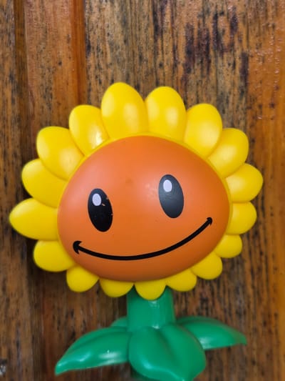 This media file features a close-up, top-down view of a toy sunflower on a wooden surface. The toy sunflower has a bright orange face with a black curved smile and two black oval eyes, each with a white oval highlight. Yellow petals surround the orange face. To the right of the sunflower's face are two bright green leaves, suggesting it's part of a larger toy plant. The background is a wooden surface with visible grain and texture. The lighting appears to be from above and slightly to the left, casting subtle shadows. There is no visible text in the image. The setting appears to be indoors, on a table or similar wooden surface. The toy's cheerful design evokes a playful or happy emotion.
